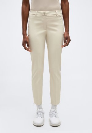 Person trägt schlanke beige Hose mit vorderer Knopfleiste und Reißverschluss, kombiniert mit weißen Socken und weißen Schnürschuhen, steht vor einfarbigem Hintergrund.