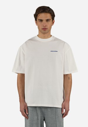 Young man wearing a white oversized T-shirt with "PEGADOR" logo on left chest and gray patterned pants against a plain background.