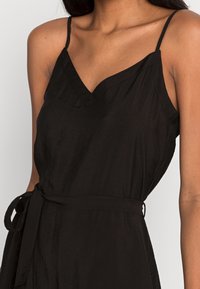 Černý overal s tenkými ramínky, véčkovým výstřihem a páskem v oblasti pasu. Tkanina má strukturovaný povrch, což dodává jemný detail tomuto designu.
