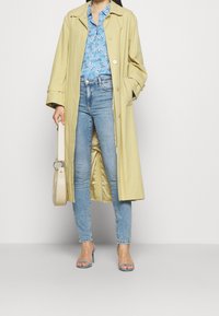 Trench coat giallo chiaro, camicia blu a fiori, jeans blu chiaro e tacchi alti con cinturino blu. Tiene una borsa a tracolla beige.