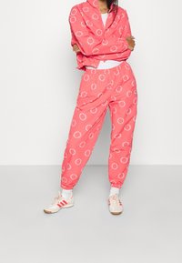 Persona che indossa una vivace tuta Adidas color corallo con motivi a logo circolari, calze bianche e sneakers beige con strisce rosse, in piedi con le braccia incrociate.