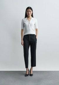 Γυναίκα που στέκεται μπροστά σε απλό γκρι τοίχο, φορώντας λευκό πάνω με κοντά μανίκια και γιακά, μαύρο ψηλόμεσο παντελόνι με κουμπιά και μαύρες ψηλοτάκουνες.