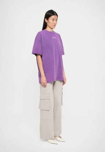 Lilla oversized t-skjorte med korte ermer, med en liten hvit logo. Parret med lysebeige cargo-bukser med sidelommer.