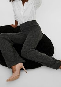 Pantalones de pierna ancha grises con cintura alta, hechos de una tela suave, combinados con tacones puntiagudos nude sobre un cojín negro texturizado. Camisa blanca de botones.