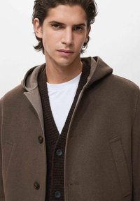 Joven con cabello oscuro que lleva un abrigo marrón con capucha sobre un cárdigan marrón oscuro abotonado y una camisa blanca, contra un fondo claro y liso.