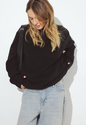 Sieviete ar blondiem matiem, tērpusies melnā džemperī un gaiši zilās džinsās, skatās uz leju ar rokām kabatās pret baltu fonu.