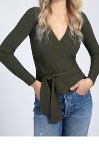 Haut cache-cœur à manches longues côtelé vert olive avec un décolleté en V profond et un détail de taille nouée, associé à un jean bleu clair déchiré.