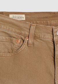 Jean Levi's Premium foncé montrant la poche avant, la boucle de ceinture, les rivets en cuivre, le bouton en métal et une étiquette partiellement visible à l'intérieur de la ceinture.