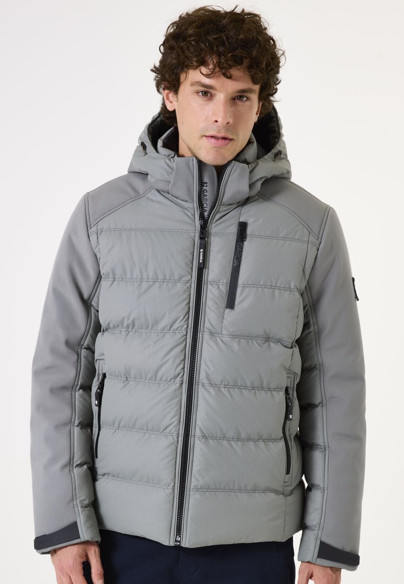 Chaqueta de abrigo gris con capucha, que presenta un tejido acolchado, bolsillos con cremallera y un detalle de cremallera negra. Material suave y aislante.