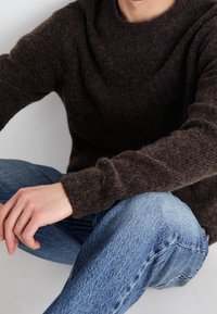 Pull en laine marron avec un col rond, porté sur un jean denim bleu clair. Le pull présente une finition texturée et des poignets côtelés.