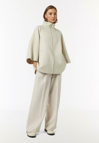 Giacca zip beige chiaro con colletto alto e maniche larghe, abbinata a pantaloni beige chiaro a vestibilità morbida e scarpe color crema. Texture liscia.