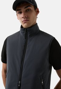 Grau gepolsterte Weste mit hochgelegenem Kragen, Frontreißverschluss und seitlichen Reißverschlusstaschen. Über einem figurbetonten schwarzen Shirt getragen. Glatte, matte Textur.