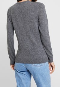 Vrouw gekleed in een nauwsluitende grijze lange mouwen trui en lichtblauwe hoge taille jeans, gezien van achteren tegen een effen achtergrond.