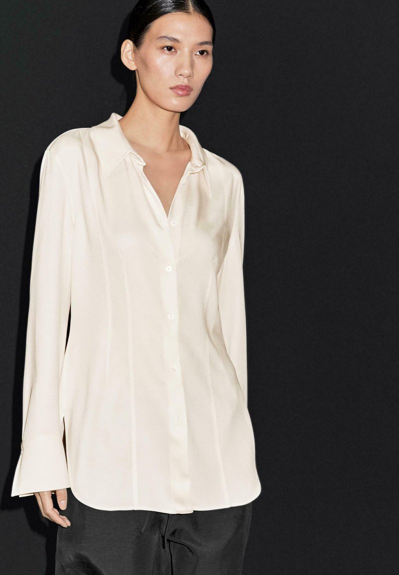 Blusa di seta beige con colletto classico, maniche lunghe e leggera lucentezza, caratterizzata da un'apertura con bottoni e una vestibilità rilassata.
