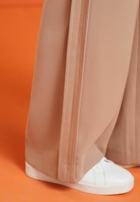 Pantalons larges en beige doux avec deux bandes blanches sur les côtés ; associés à des baskets blanches. Fond orange. Texture de tissu lisse.