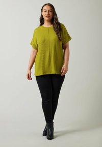 Camiseta ligera de color verde oliva con mangas cortas, que presenta un pliegue central y un corte holgado. Combinada con jeans skinny negros y botas de tobillo.