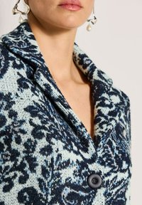 Frau trägt einen blau-weiß gemusterten Strickblazer mit großem schwarzen Knopf und herabhängenden Perlenohrringen, Lippen geschlossen, neutraler Hintergrund.