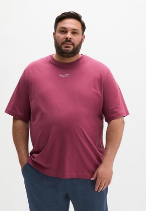 Homme barbu portant un t-shirt Marc O'Polo rose foncé et un pantalon bleu marine, debout devant un fond uni.