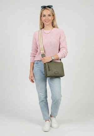 Vrouw met lang blond haar die een roze trui draagt, lichtblauwe spijkerbroek, witte sneakers en een olijfgroene crossbodytas met een gevlochten riem draagt.