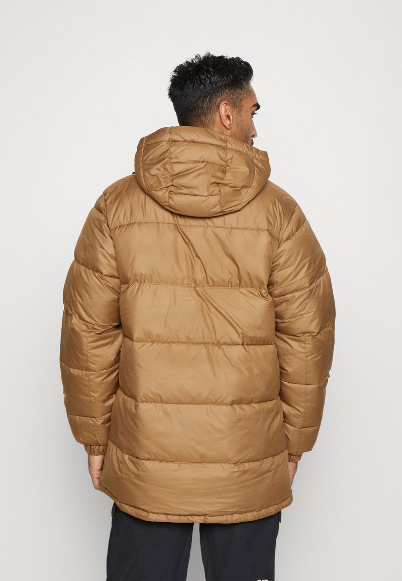 Mężczyzna widziany z tyłu, ubrany w brązową ocieplaną kurtkę z kapturem i czarne spodnie na jednolitym jasnym tle.