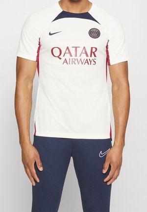 Homme portant un maillot de football blanc du Paris Saint-Germain avec le logo Qatar Airways et un pantalon Nike bleu marine sur fond uni.