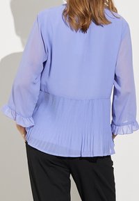 Ljuslila blus med genomskinliga långa ärmar, plisserad bakdel och volangärmar, ihop med svarta byxor. Slät textur, avslappnad passform.