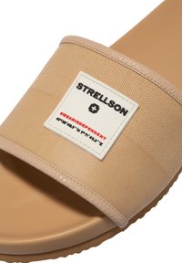 Béžové skládací sandály s texturovaným svrškem, hladkým lemem a obdélníkovým logem s černým a červeným textem. Gumová podrážka se vzorkem proti skluzu.