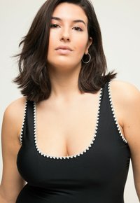 Černé plavky s hlubokým výstřihem a bílým trojúhelníkovým lemem podél okrajů. Hladká tkanina se ramínky a zakřivenou siluetou.