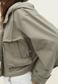 Vrouw in een lichtolijfgroene hooded jack met een frontale rits en grote ritszak, gecombineerd met witte broek.