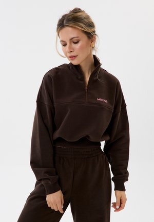 Frau trägt dunkelbraunen Zip-up-Sweatshirt und passende Hose, schaut nach unten, Haare zurückgebunden und goldene quadratische Ohrringe.