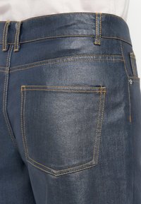 Jean en denim bleu foncé avec des coutures jaunes, présentant une poche arrière et une texture lisse. La taille est également mise en valeur.