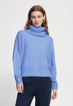 Jeune femme aux longs cheveux bruns portant des boucles d'oreilles créoles en argent et un pull à col roulé bleu clair avec des poignets boutonnés et un pantalon foncé.