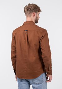 Camisa marrón de manga larga con cuello estándar, textura suave y dobladillo redondeado. Cuenta con un pliegue en la parte trasera y puños abotonados.