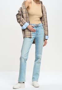 Jeans de un azul claro, top beige sin mangas y una camisa de cuadros marrón con puños azules. Cinturón plateado con iniciales. Zapatillas blancas completan el atuendo.