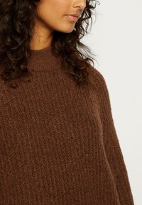 Pull en maille côtelée marron avec un col montant, doté d'une texture douce et d'une coupe décontractée. Coutures visibles et détails de motif subtils.