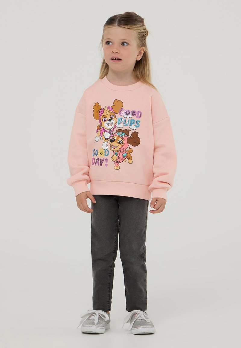 Felpa rosa chiaro con illustrazioni di cani giocosi e la scritta "Buona giornata!", abbinata a jeans grigi e sneaker argentate.