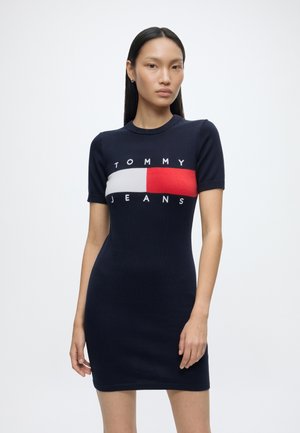 Femme aux longs cheveux foncés portant une robe bleu marine ajustée à manches courtes avec un logo Tommy Jeans rouge, blanc et bleu marine sur la poitrine.