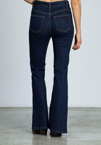 Jeans a campana blu scuro con vita alta, cinque tasche, cuciture visibili e una texture in denim liscia. Indossati con stivali con tacco marroni.