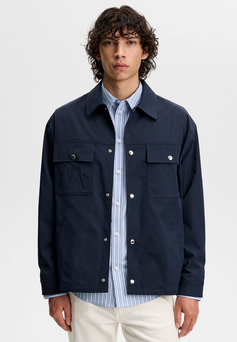 Giovane uomo con capelli ricci che indossa una giacca blu a bottoni sopra una camicia a righe blu e pantaloni chiari, in piedi davanti a uno sfondo semplice.