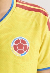 Gul fodboldtrøje med røde og blå stribede accenter, der har et rundt logo med et rødt hexagon-mønster og "Federación Colombiana de Fútbol."
