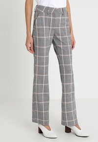 Vrouw in grijs en roze geblokte rechte broek, gecombineerd met witte blokhakken en een witte top, staande tegen een effen achtergrond.