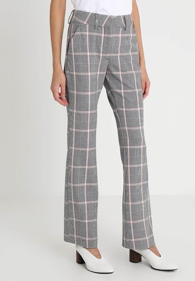 Vrouw in grijs en roze geblokte rechte broek, gecombineerd met witte blokhakken en een witte top, staande tegen een effen achtergrond.