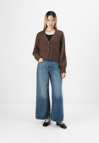 Kvinne iført brun knappet strikkegenser, svart topp, brede blå jeans og svarte balettleiligheter som står mot hvit bakgrunn.