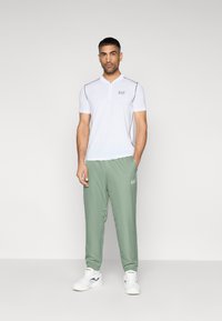 Λευκό πόλο με κοντά μανίκια, μαύρες λεπτομέρειες και λογότυπο, σε συνδυασμό με ανοιχτοπράσινο παντελόνι και λευκά αθλητικά παπούτσια με μπλε διακοσμητικά στοιχεία.