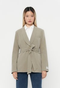 Světle šedý blazer s proužky, s páskem na zavazování, klopovým límcem a jednou náprsní kapsou, nošený přes bílou košili a modré džíny.