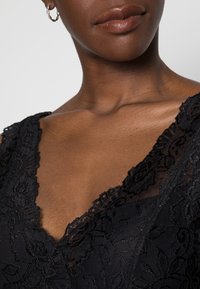 Indumento in pizzo nero con design a V; presenta motivi floreali e un delicato pizzo lungo il colletto, con una texture morbida.
