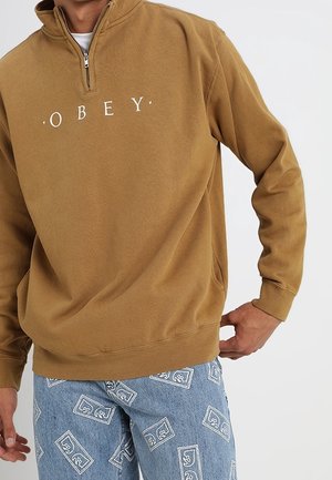 Mann trägt ein braunes Sweatshirt mit Viertel-Reißverschluss und dem Aufdruck "OBEY" sowie blaue Jeans mit weißen rechteckigen Logos.