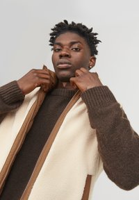 Beige and brown fleece jacket with a zip front, ribbed cuffs, and a slight sheen. Texture varies between fleece and smooth fabric.