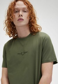 Camiseta de algodón verde oliva con mangas cortas y cuello redondo. Presenta un logo bordado de "FRED PERRY" en verde más oscuro en el pecho.