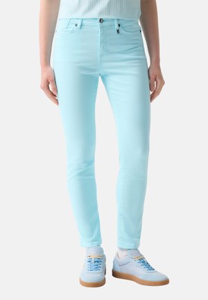 Persoon die lichtblauwe skinny jeans, witte sokken en witte sneakers met lichtblauwe veters draagt, staand tegen een effen achtergrond.
